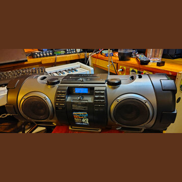 Šedý přenosný radiomagnetofon značky JVC. Na předním panelu je digitální displej, ovládací tlačítka a nápis POWERED BOOMBOX CD SYSTEM RV-KN401. Ve sp…