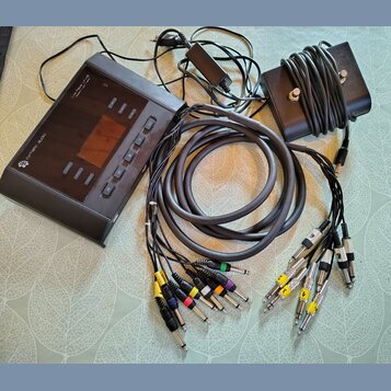 Černý elektronický přístroj s nápisy "CYMBIC AUDIO" a "Live Pipe LP 16". Součástí je černý napájecí adaptér, černý kabel svázaný na hranatém podložko…