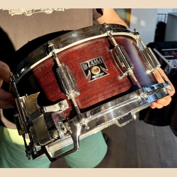 Červený dřevěný snare drum s kovovými obručemi a mechanismem na straně. Na korpusu je čitelný nápis TAMA SUPERSTAR a pod ním text MADE IN JAPAN. Povr…
