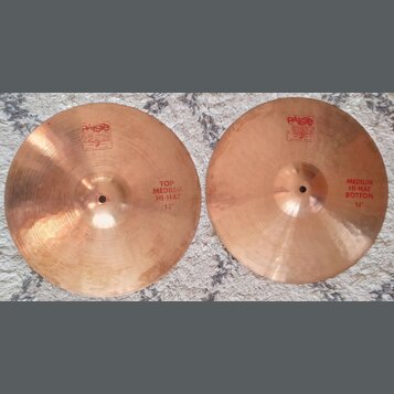 Dva mosazné činely typu hi-hat položené na šedém koberci. Levý činel nese nápisy „PAISTE 2002“, „TRADE MARK MADE IN SWITZERLAND“ a „TOP MEDIUM HI-HAT…