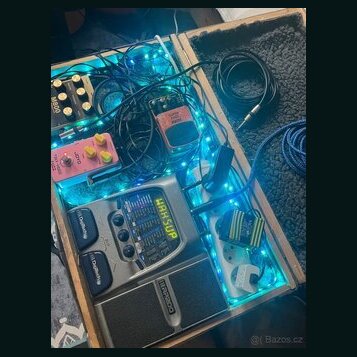 Dřevěný kufr s pedalboardem, který je podsvícen modrými LED pásky. Obsahuje efektové pedály a procesor s nápisy „BOSS“, „MB-800“ a „MAH SUP“. V kufrě…