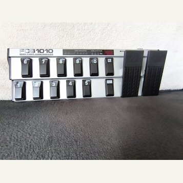 Šedý MIDI kontrolér s nápisem FCB1010 MIDI FOOT CONTROLLER. Zařízení obsahuje dvě řady černých spínacích pedálů s bílými čísly 1 až 10 a tlačítky UP…