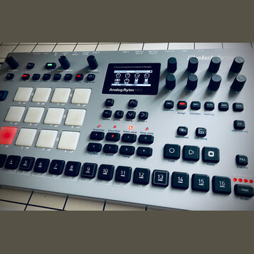Stříbrný digitální drum machine s názvem Analog Rytm MKII a logem Elektron. Zařízení obsahuje displej, zahlreiche ovládací knofíky, bílé gumové pady…