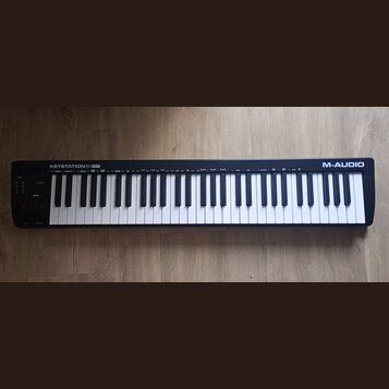 Černá klaviatura s bílými a černými klávesami. Na pravém horním okraji je nápis KEYSTATION 61 MK3 a v pravém dolním rohu logo M-AUDIO. Povrch nástroj…
