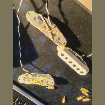 Tři elektrické snímače kytary v krémové barvě. Na jednom je nápis Suhr a 2,1H, na druhém SuhrV70 a 2,4H. Snímače jsou omotány žlutými gumičkami a ved…