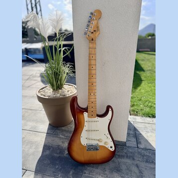 Elektrická kytara s trupem v barevnosti sunburst a hnedým hmatníkem. Na hlavě je viditelné logo Fender. Kytara má bílý pickguard, tři snímače a kovov…