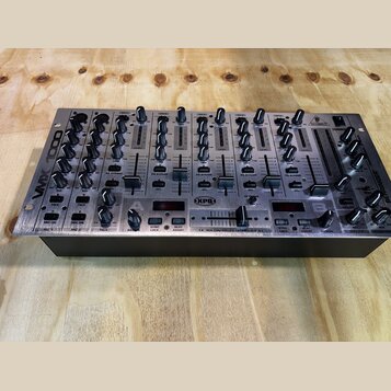 Stříbrný DJ mixér s černými ovladači a posuvníky. Na levém boku je nápis VMX 1000 a na pravém boku logo BEHRINGER. Uprostřed spodní části je nápis XP…