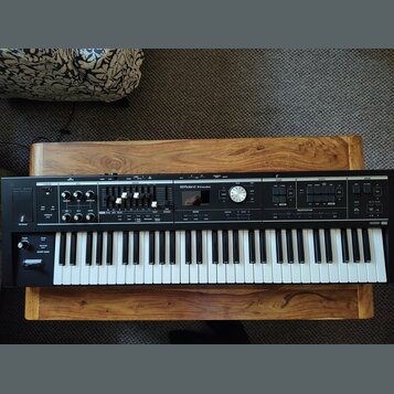 Černý digitální klavír s bílými a černými klávesami, umístěný na dřevěném stole. Na ovládacím panelu je nápis Roland V-Combo VR-09. Přístroj obsahuje…