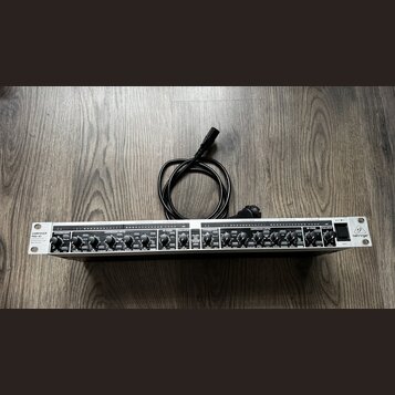 Stříbrný rackový procesor s černými ovládacími prvky a nápisem COMPOSER PRO-XL. Na pravé straně je logo behringer a vypínač POWER. Za přístrojem leží…