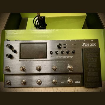 Stříbrný kytarový procesor s nápisem GE 300 a doplňkem Amp modelling & Stomp & Multi Effects. Zařízení má displej, několik otočných ovladačů a řadu s…