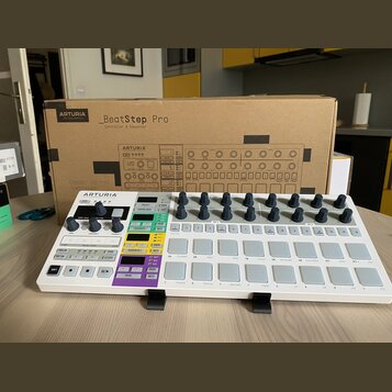 Bílý sekvencer Arturia BeatStep Pro umístěný na černém stojanu před svou originální kartonovou krabicí. Na přístroji jsou vidět šeradovky šedých pods…