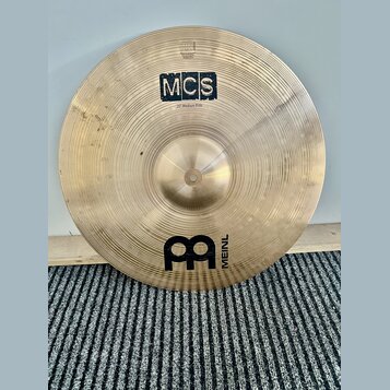 Zlatavý kovový činel s viditelným broušením. Na povrchu jsou nápisy MCS a 20" Medium Ride, v dolní části je logo MEINL. V horní části je malý nečitel…