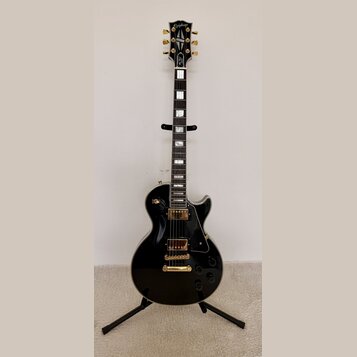 Černá elektrická kytara s gold hardwarem umístěná na černém stojanu. Na hlavě hmatníku je bílý nápis Epiphone. Kytara má dva humbuckery, zlatý most a…