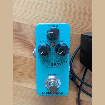 Modrý kytarový efekt s nápisy HYPERGRAVITY a MINI COMPRESSOR. Na horní straně jsou tři černé otočné ovladače s popisky SUSTAIN, ATTACK a LEVEL. Ve sp…