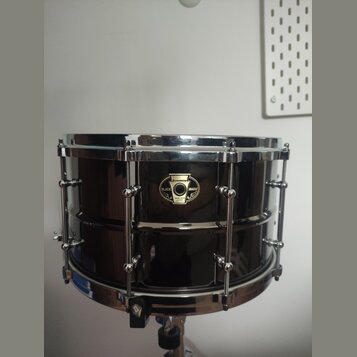 Černý malý snare drum s chromovaným kováním. Na koruse je umístěn oválný štítek s nápisy „pawpaw“, „BLACK MAGIC“ a „SNARE DRUM LIMITED“. Povrch nástr…
