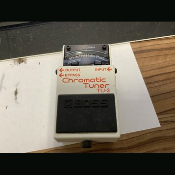 Bílý kytarový ladič značky BOSS, model TU-3 Chromatic Tuner. Na horní straně je umístěn displej s analogovou stupnicou a nápisem CHECK. V dolní části…