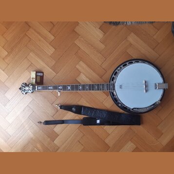 Na fotografii je banjo s bílou membránou a kovovým tělem, k němuž je přiložen černý popruh a malá krabička s příslušením. Na hmatníku jsou vidět orna…