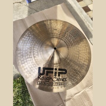 Zlatavý kovový činel s kladivkovaným povrchem, položený na hnědém kartonu. Na povrchu je černý nápis UFIP Rough Series EARCREATED CYMBALS a malá nále…