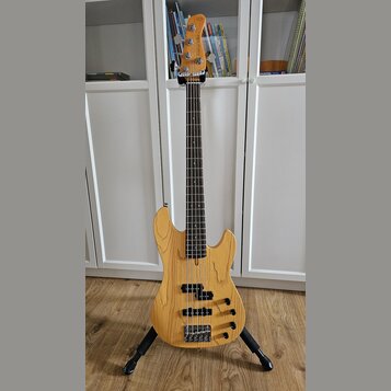 Pětstrunná basová kytara s přírodním dřevěným povrchem těla a hmatníkem z tmavého dřeva. Na hlavě nástroje je svisle napsáno „Marcus Miller Deluxe 5“…