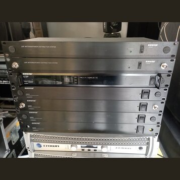 Snímek zachicuje stoh rackových zařízení. V horní části jsou dvě šedé jednotky s nápisem UHF ANTENNA/POWER DISTRIBUTION SYSTEM a logem SHURE UA844. P…