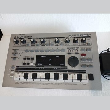 Stříbrný sekvenční syntezátor Roland MC-303 groovebox. Na předním panelu jsou umístěny ovládací knofíky, tlačítka a displej s nápisy jako CURRENT, NE…