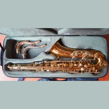 V otevřeném kufru značky Bam leží zlatý saxofon a dva zakřivené hrdly s korkem. Nástroj má lesklý povrch a je vybaven řadou klapek a mechanismů. Na s…