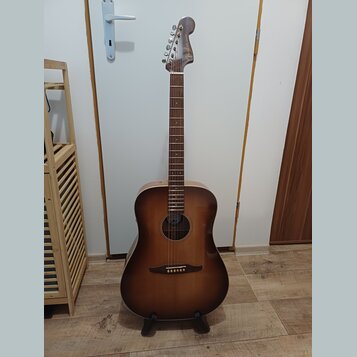 Akustická kytara s korpusem v tmavě hnědém přechodovém odstínu (sunburst) a hnedým hmatníkem. Na hlavě nástroje je viditelné logo Fender. Kytara stoj…