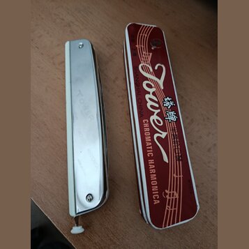 Na fotografii je chromovaná chromatická harmonika a její červené kovové pouzdro. Na nástroji je vyražen nápis TOWER, CHROMATIC HARMONICA a MADE IN CH…