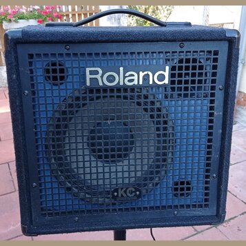 Černý kombozesilovač s povrchem z textilie a kovovou ochrannou mřížkou. Na mřížce je nápis Roland a v dolní části logo KC s doplňkem KEYBOARD AMPLIFI…