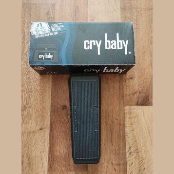 Na fotografii je černý wah pedál s gumovou texturovanou povrchovou vrstvou, umístěný před svou originální krabicí. Na krabici je bílý nápis cry baby.…