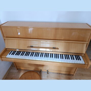 Světle hnědé pianino s lakovaným dřevěným povrchem. Na krytu nad klávesnicí je viditelný nápis "SMILE". Nástroj vykazuje známky opotřebení v podobě š…