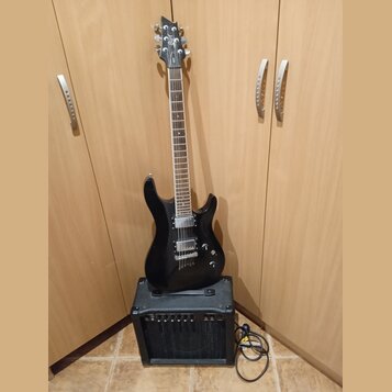 Černá elektrická kytara s dvojstupňovými humbuckery, opřená o černý kytarový kombo zesilovač. Na hlavě kytary je bílý nápis Cort. Zesilovač má na pře…