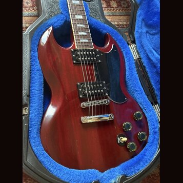 Kytara s červeným lakem a černým plechtrátem, umístěná v otevřeném kufru s modrou výplní. Nástroj má dva černé humbuckery, chromovaný most a čtyři če…