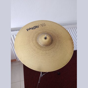 Zlatý kovový činel umístěný na stojanu. Na povrchu je černý nápis PAiSte 302 a pod ním menší text 20" Ride. Povrch činelu vykazuje drobné škrábance a…