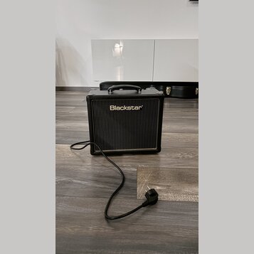 Černý kytarový kombo zesilovač s rukojetí nahoře a černou síťovkou. Na přední straně je bílý nápis Blackstar®. Z přístroje vede napájecí kabel s vidl…