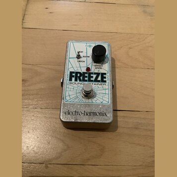 Kytarový efekt pedál v kovovém šasi s bílo-modrým grafickým potiskem. Na horní straně je nápis FREEZE, pod ním text SOUND TAINER a ve spodní části lo…