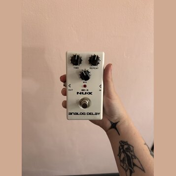 Bílý kytarový efekt s nápisy NUX, ad-3 a ANALOG DELAY. Na horní straně jsou tři černé otočné ovnovky s popisky TIME, REPEAT a MIX. V dolní části je k…