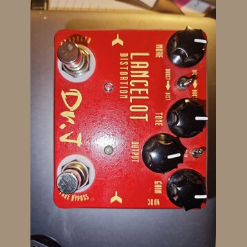 Červený kytarový efekt s nápisy LANCELOT DISTORTION a D.T. na horní straně jsou čtyři černé otočné knoflíky s popisky MORE, TONE, OUTPUT a GAIN. Na p…