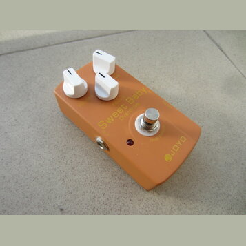 Oranžový kytarový efekt s bílými oválovými knoflíky. Na horní straně jsou nápisy Volume, Focus, Sweet Baby, Overdrive, OUT, 9V DC a TRUE BYPASS, v pr…