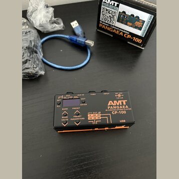 Černý kompaktní efekt pedál s oranžovým spodním okrajem. Na horní straně je nápis AMT PANGAEA IR CONVOLUTION PLAYER CP-100, malý displej a ovládací t…