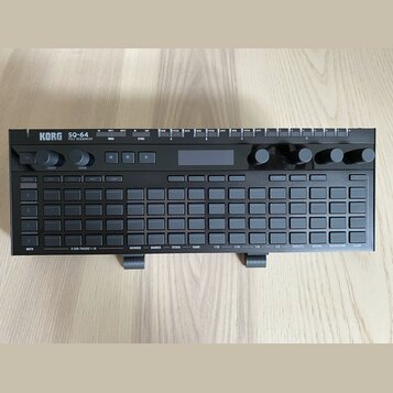 Černý polysequencer s nápisy KORG a SQ-64 POLY SEQUENCER. Zařízení obsahuje mřížku šedých tlačítek, několik otočných ovladačů a malý displej. Na horn…