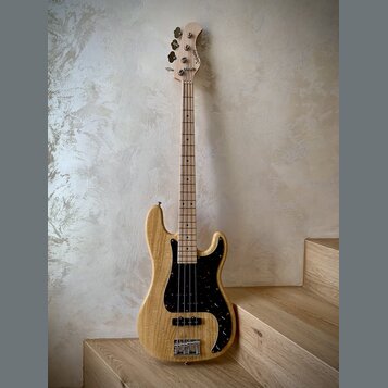 Elektrická basová kytara s přírodním dřevěným povrchem těla a hmatníku. Nástroj má černý pickguard s bílými tečkami, dva snímače a čtyři ladičky. Na…