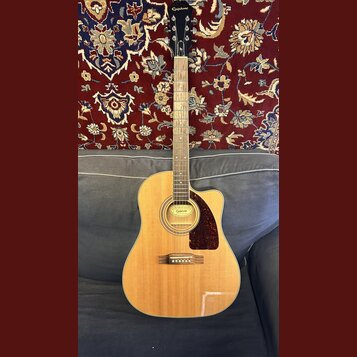 Akustická kytara s vyřezem v těle, přírodní dřevěnou barvou a hnědým hmatníkem. Na hlavě je bílý nápis Epiphone a na štítku uvnitř otvoru zvukové dír…