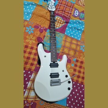 Bílá elektrická kytara s tmavým hnedým hmatníkem. Na hlavě je viditelné logo MUSIC MAN. Kytara má dva humbuckery, tremolo s páčkou a tři ovládací kno…