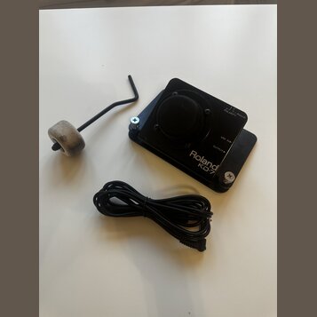 Černý elektronický trigger Roland KD-7 s připojeným kabelem. Na horní části je přepínač POLARITY a vstupy MIX IN a OUTPUT. V doprovodu je imbusový kl…