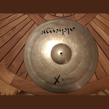 Kovový činel v přirozené zlatavé barvě s viditelným rýhováním. V horní části je černý nápis „alchemy cymbals“. Ve spodní části je černý kříž a vedle…