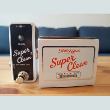 Bílá efekt pedál s černým předním panelem a kovovým spínacím tlačítkem. Na panelu jsou nápisy GAIN, Super Clean a BY xotic usa. Vedle pedálu je bílá…