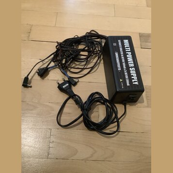 Černý napájecí adaptér s nápisem MULTI POWER SUPPLY. Na boku je uveden text SEPARATE ISOLATED 6x DC10V/100mA & 2x DC10V/200mA - SMRCKAEFFECTS - a ozn…