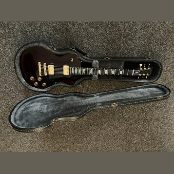 Tmavě hnědá elektrická kytara s bílým ráfkem v černém tvrdém kufru. Na hlavě kytary je viditelné logo Gibson. Nástroj má dva humbuckery, zlatý hardwa…