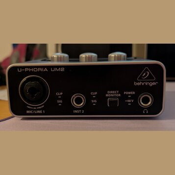 Černý zvukový interface s kovovým stříbrným rámem. Na přední straně jsou nápisy U-PHORIA UM2 a logo behringer, vstup MIC/LINE 1 s nápisem XENYX MIC P…